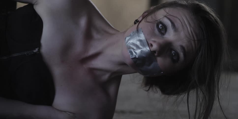 A woman in a room on the floor with her mouth duct taped with a wall in the background