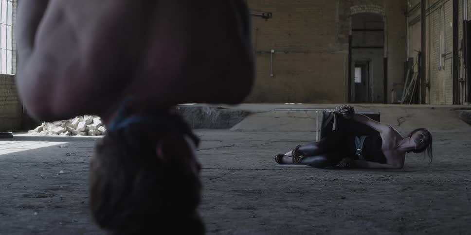 A woman tied to a chair with her mouth taped shut struggles on the floor of a building as a man hangs upside down in front of her with this hands tied together