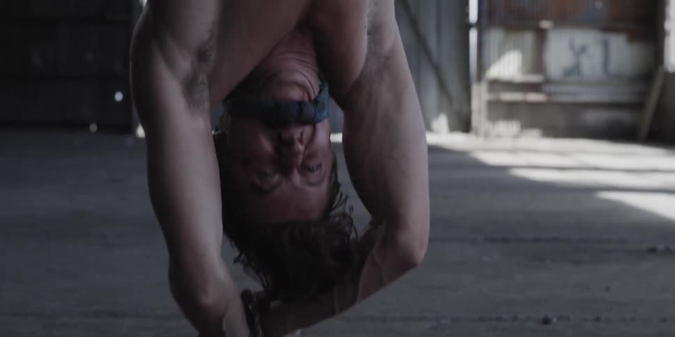 Nervous shirtless man tied up hanging upside down inside a warehouse