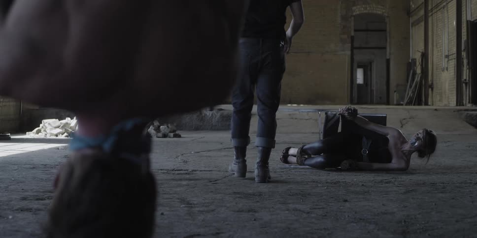 A man with a weapon walks toward a woman who is bound and gagged on the floor