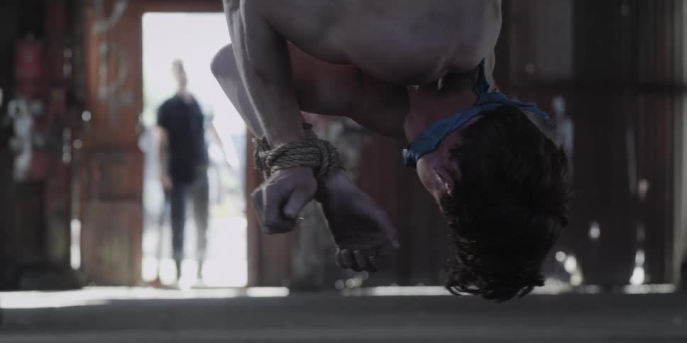 A tied up man hangs upside down in a warehouse as another man walks in with an ax
