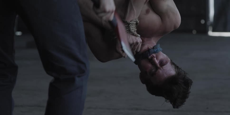 Man holding ax crouches to face man hanging upside down gagged and bound