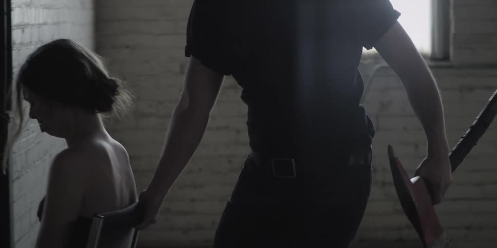 A man holding an axe pulls a chair with a woman tied to it and gagged across a room