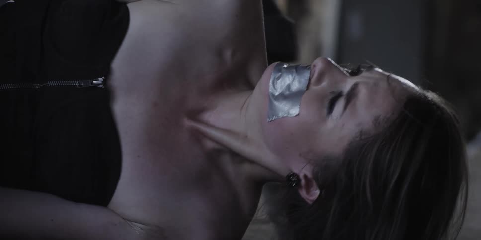 A woman on the floor with duct tape on her mouth with a person with boots picking her up off the floor in a room with a wall in the background