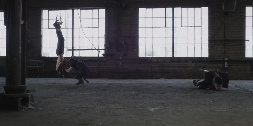 Man menacingly threats a man hanging upside down near a woman tied to a chair inside a warehouse