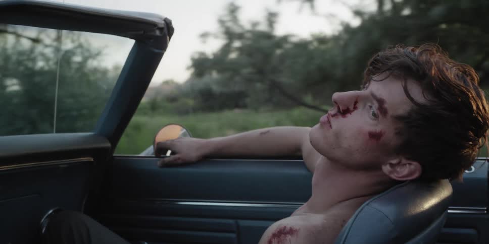 A wounded man sits in the passenger seat of a vehicle as it travels down a countryside road