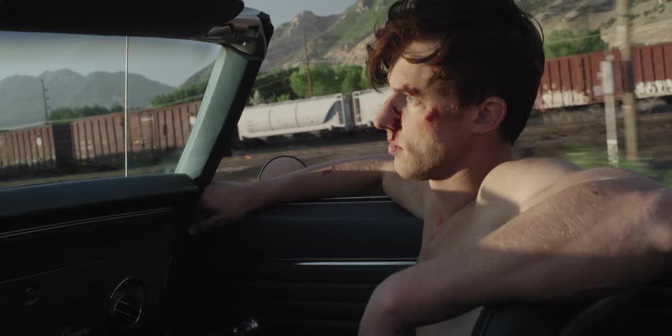 A man riding in a vehicle with bruises on his face with trains a mountain range the sky and clouds in the background