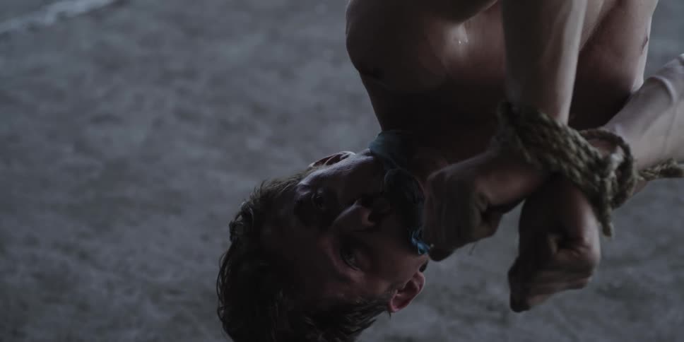 A man hanging upside down with his hands tied with a rope with another man standing over him