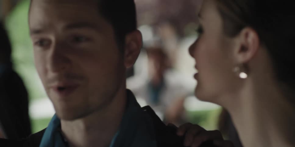 A man sits on a motorcycle and watches a woman who looks at him and then kisses the man next to her