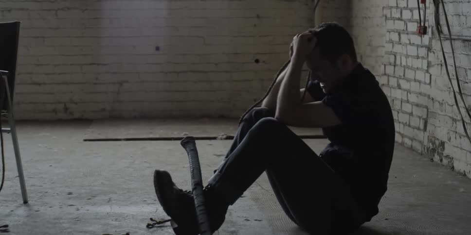 A man sitting on a floor in a room kicks a chair in anger
