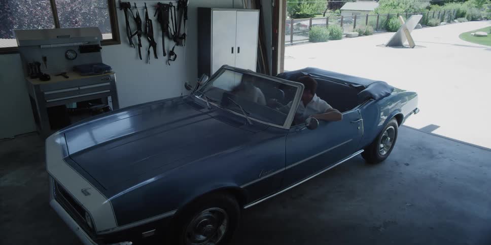 A man and woman enter a parked car and pull out of a garage on a sunny day