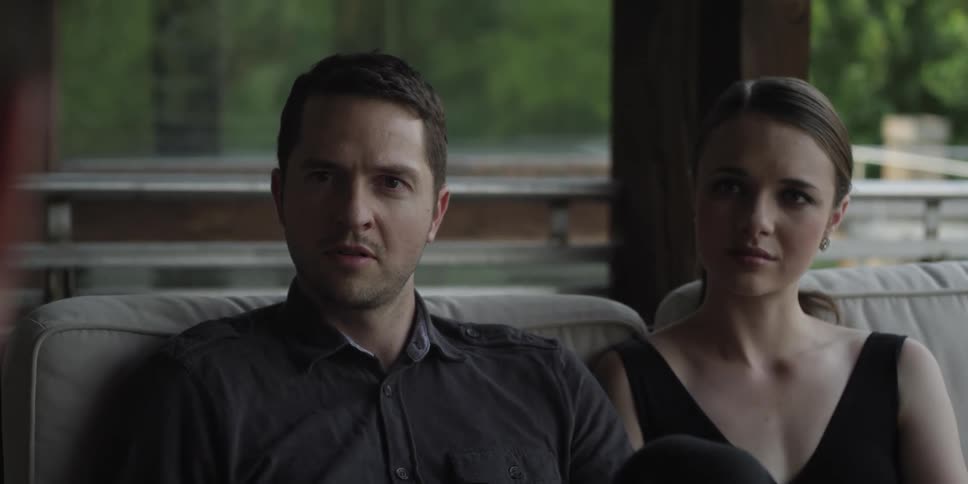Two men and a woman in a room sitting on a couch with a wall and window in the background