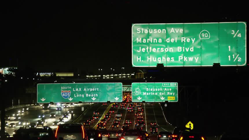 A highway with vehicles signs and streetlights with buildings in the background