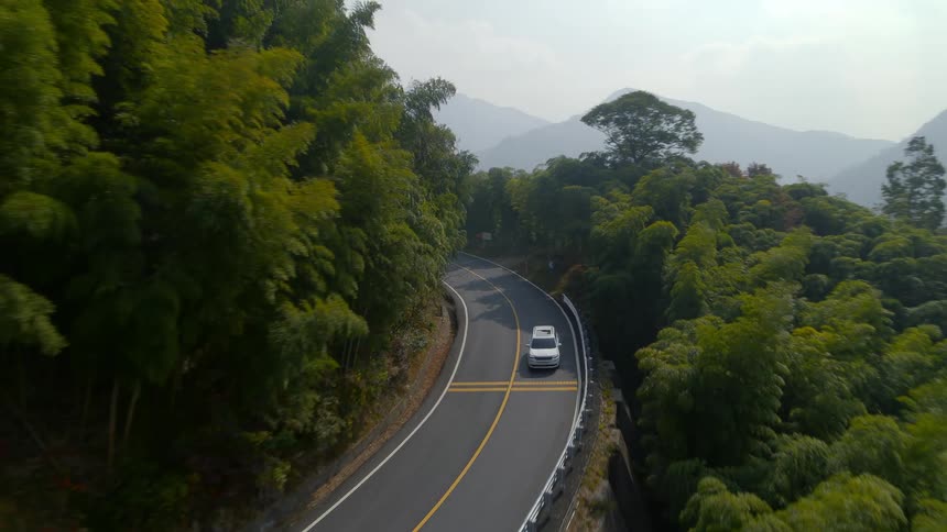 Aerial view of a white van traveling on a road surrounded by trees and mountains in the distance
