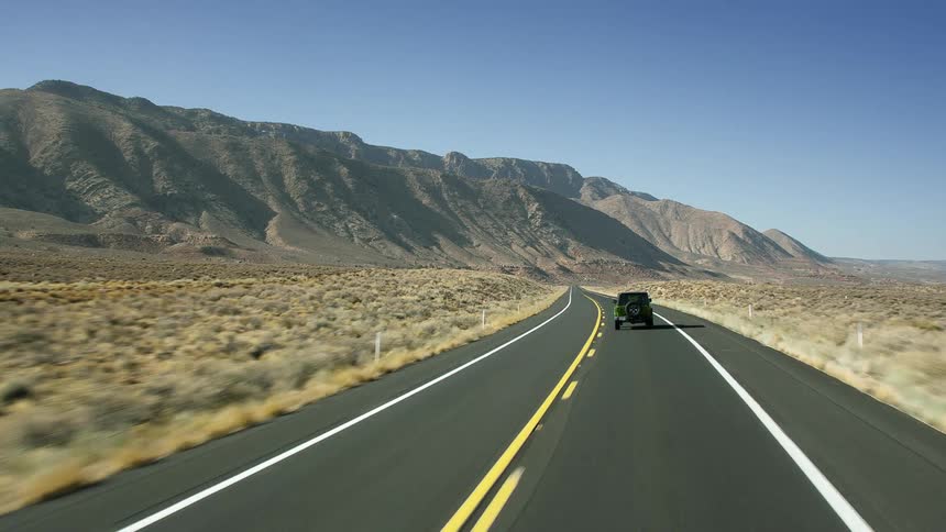 A jeep is traveling on a road towards mountains under a blue sky