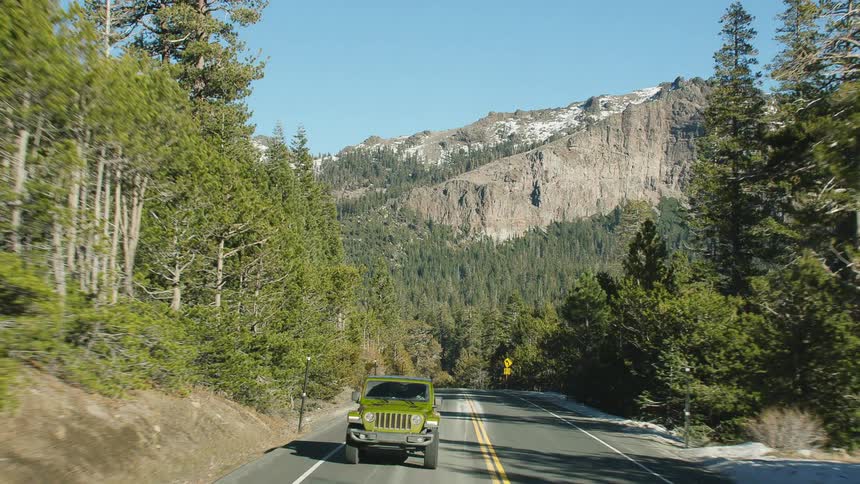 A green jeep travels on a road surrounded by trees and a mountain range is in the distance