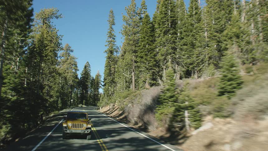 A jeep drives down a smooth 2 lane road surrounded by pine trees