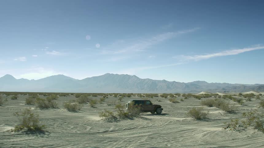 Jeep drives across desert paralleling mist covered mountains under lightly cloudy sky