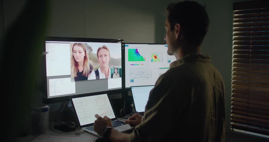 A man at his desk with two laptops and two screens in front of him waves at a man and woman he is video chatting with