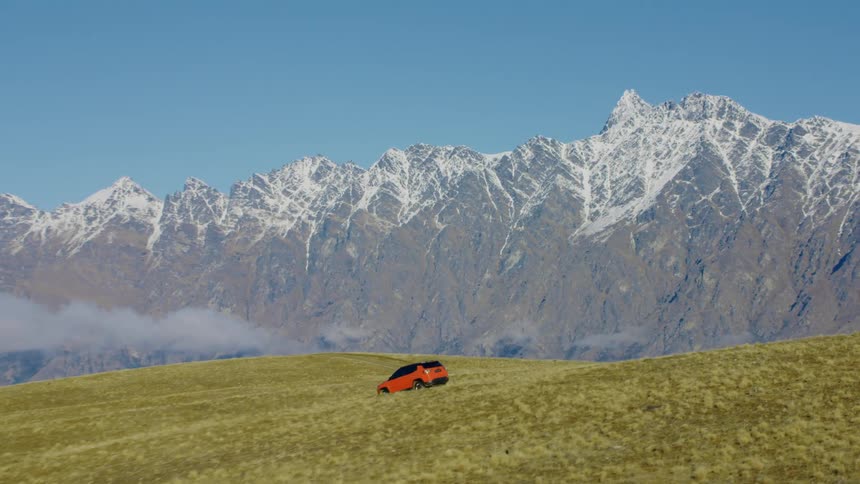 A red suv bumps along green hills opposite an icy mountain ridge