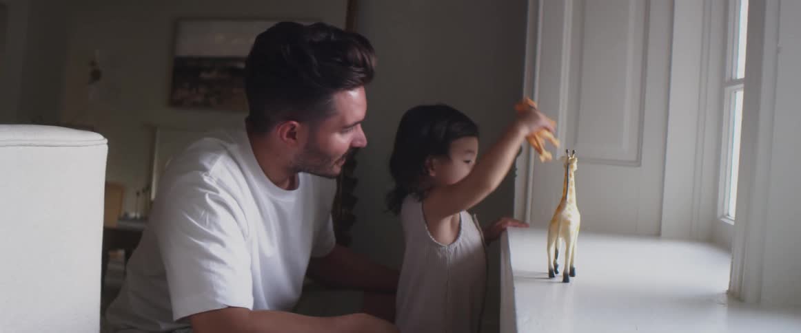 A father watches his child play with toy animals on a window sill at home