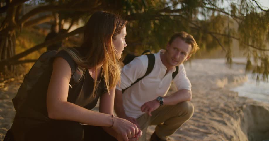 Young couple crouching under the trees on the coast near the beach at sunset