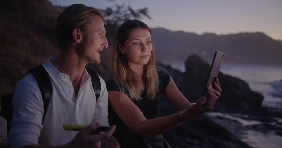 Two people using a tablet and relaxing on a coastal area