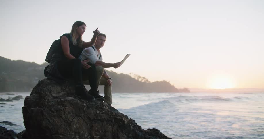 A man and woman sit on a rock on the coast and look at a piece of paper