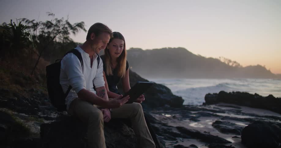 A man and woman sitting side by side on a rock near the ocean looking at a tablet