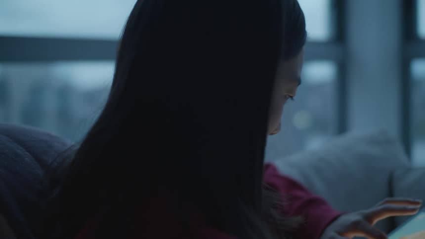 A girl sits holding a tablet and playing a game near large windows on a gray cloudy day