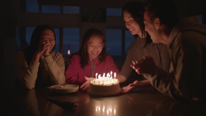 A group of people are sitting at a table as a woman brings a birthday cake over with the candles lit