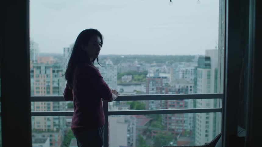 A woman stands on her balcony with the city skyline across from her and looks back in to her home