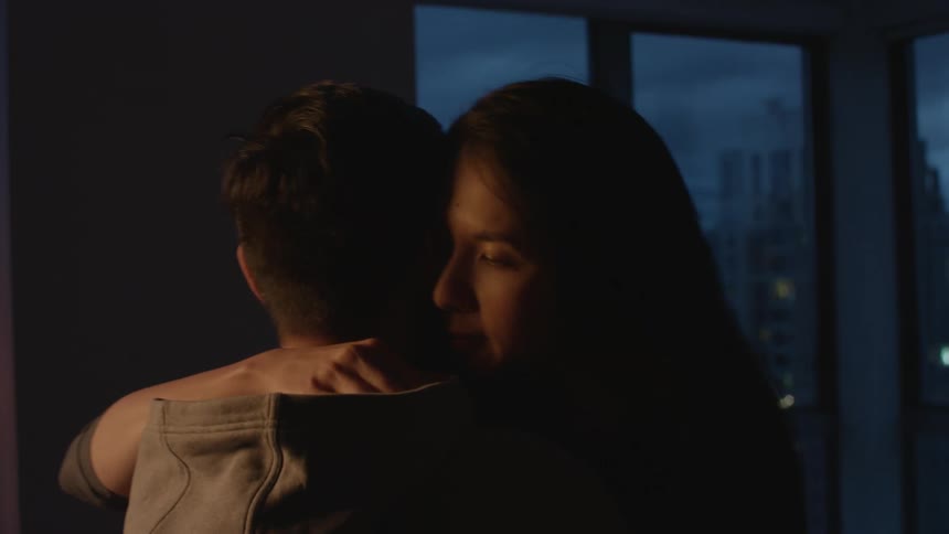 A man and a woman in a room hugging with a wall and window in the background