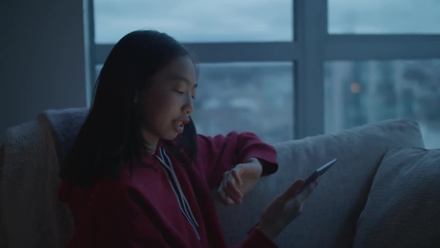 Teenage girl with straight hair and red zip up sits on grey couch and uses tablet making video call