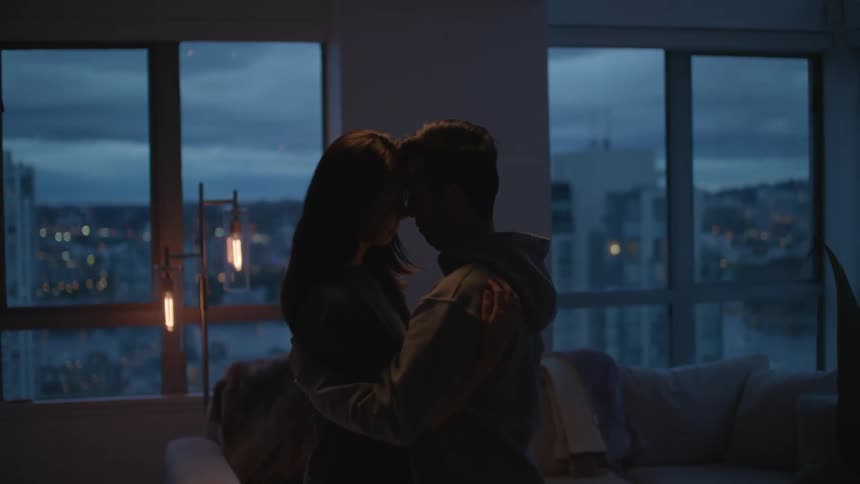 Couple slow dance in living room partly silhouetted by light from window with view of overcast sky and city after dusk