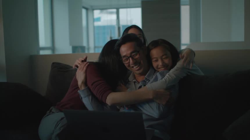 A man and a woman and two girls in a room hugging and looking at a laptop with a wall and window in the background