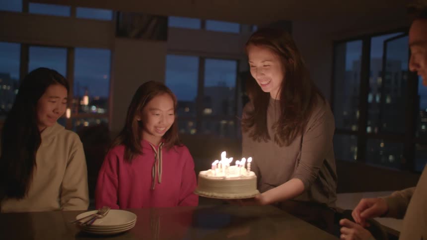 A woman surprises her daughter with a birthday cake and lit candles as the family sits at a table at home