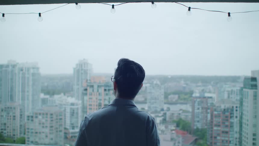 A man stands on a balcony and looks out at the city skyline