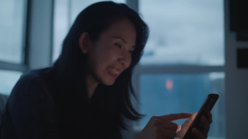 A woman laughs while using her smartphone to participate in a video chat as she sits indoors in the evening