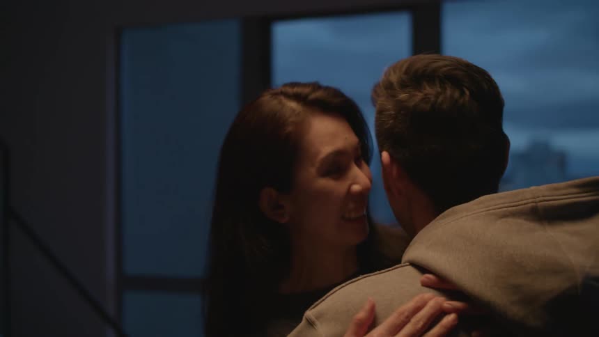A man and a woman in a room hugging with a wall and window in the background