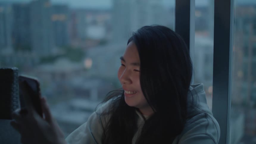A woman smiles as she holds her phone up to the window to show the cityscape below to her video call