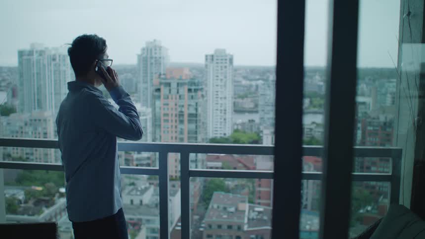 Man standing on an apartment balcony answer the phone while looking at the city