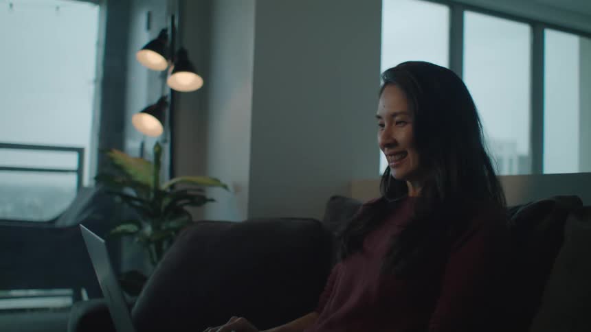 A woman smiles and laughs while participating in a video chat using a laptop while sitting on a sofa