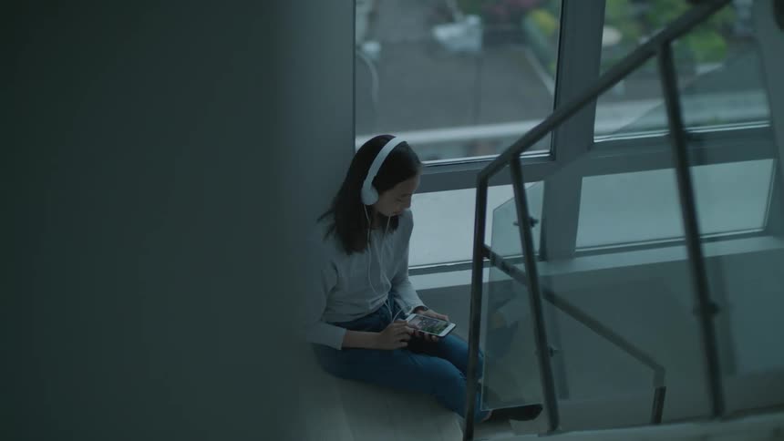 A woman is listening to music through headphones as she sits in a stairwell and looks at her phone