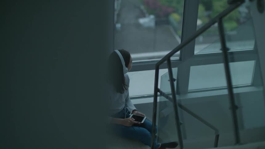 A young girl sits in a staircase and listens to music through headphones on her cell phone as she looks out the window