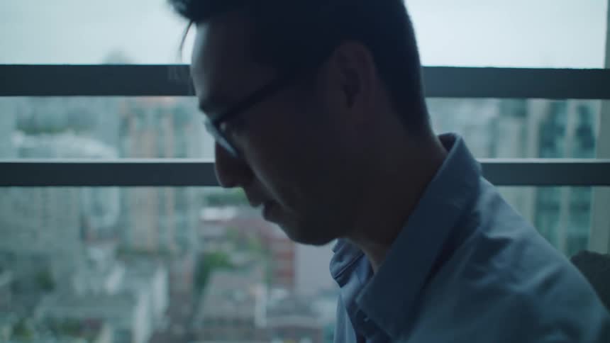 A man with glasses on a balcony using a laptop with the sky and clouds in the background