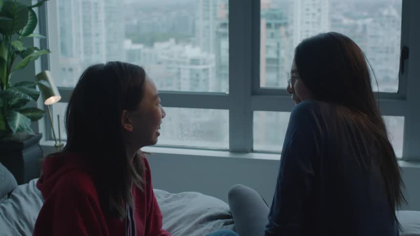 Two sisters laugh as they sit on a bed in a room with views of downtown