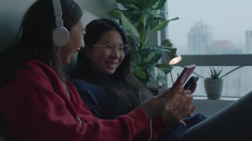 Two sisters laugh while looking at their smartphones while sitting in bed