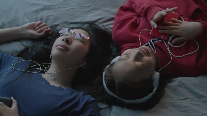 Sisters wearing earbuds and headphones listen to music while laying in bed