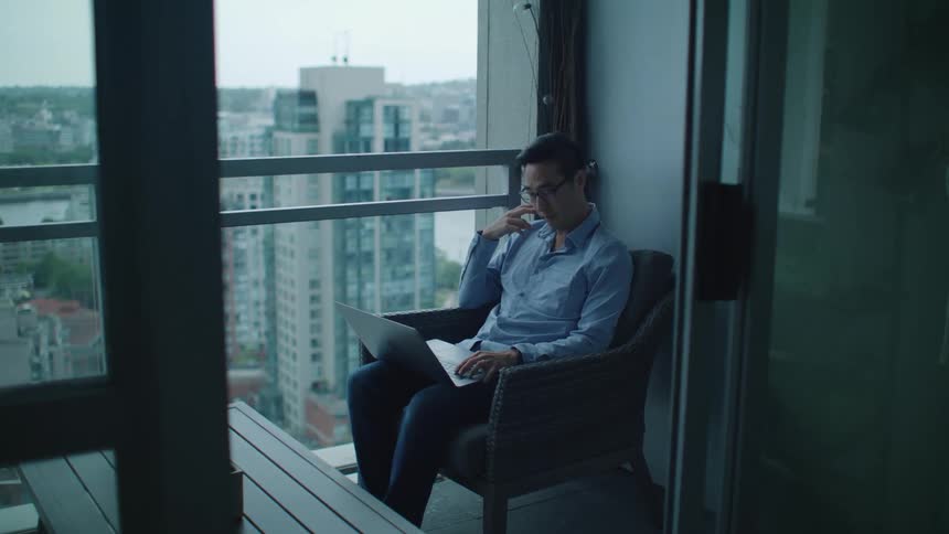 A man sits on his balcony at home with his laptop and nod his head and talks as he looks at the screen